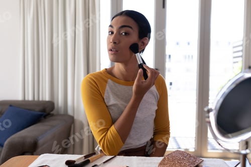 Preview: Mixed race gender fluid man putting on foundation powder with makeup brush