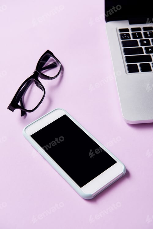 Preview: close-up shot of smartphone with eyeglasses and laptop at workplace on pink surface