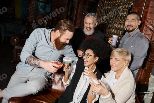 Preview: bearded tattooed man clinking glasses with cheerful women near multiethnic friends in cocktail bar