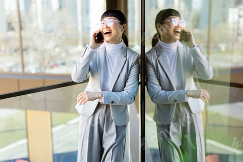 Preview: Professional Asian woman enjoys a joyful phone call while standing by a glass railing on a sunny day