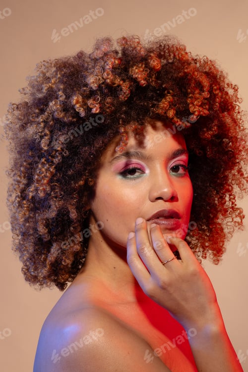 Preview: Biracial woman with curly hair wearing pink eyeshadow and lipstick touching face, copy space