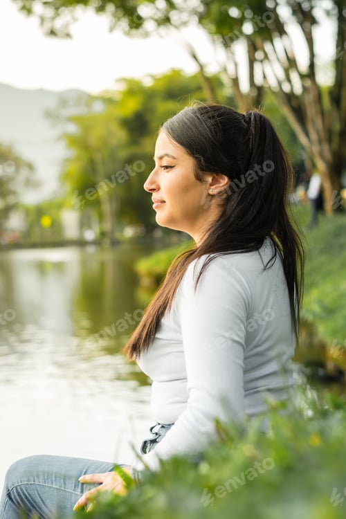 Preview: vertical shot of beautiful young latin woman looking back, sitting by a large lake