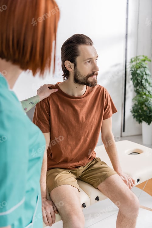 Preview: blurred physiotherapist calming displeased bearded man sitting on massage table in consulting room