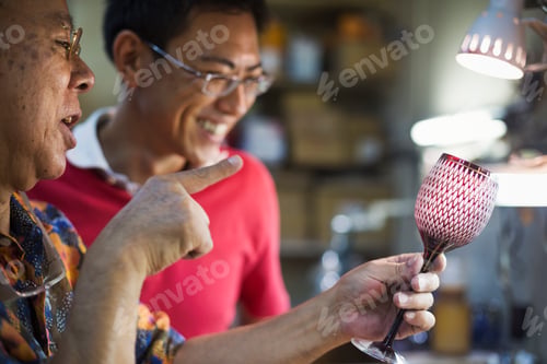 Preview: Two peoplein a glass maker's studio workshop, inspecting a red cut glass wine glass.
