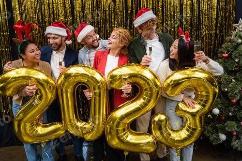 Visualização: Empresários multiculturais positivos em chapéus de Papai Noel segurando champanhe perto de balões em forma