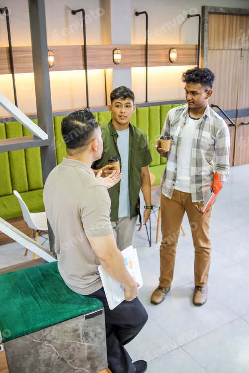 Preview: Three Male Colleagues Stand and Talk in Modern Office, Holding Papers and Coffee Cups During Break