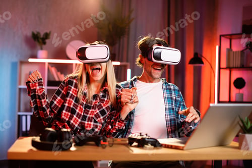 Preview: Young woman and handsome man sitting at desk and holding hands while wearing virtual reality headset