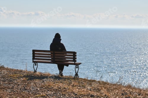 Preview: Single girl in a black jacket and hat sitting on bench at cliff at front of sea peaceful quiet place