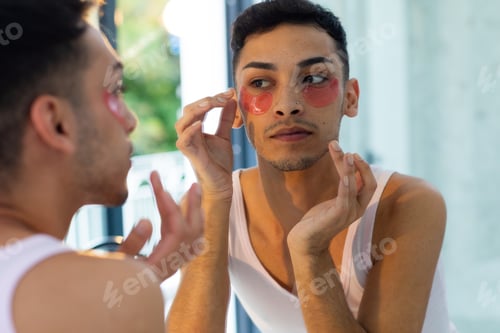 Preview: Biracial transgender man looking in mirror and applying under-eye patches in bathroom