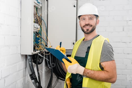 Preview: smiling handsome electrician holding clipboard near electrical box in corridor and looking at camera