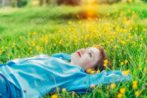 Preview: happy young woman lays on a green grass with yellow flowers, looks up and smiles at susnset