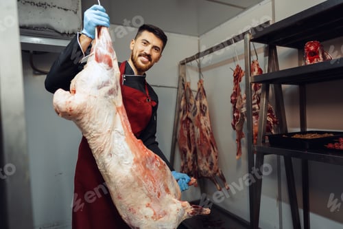 Preview: Male butcher standing by meat hanging on hook in meat shop