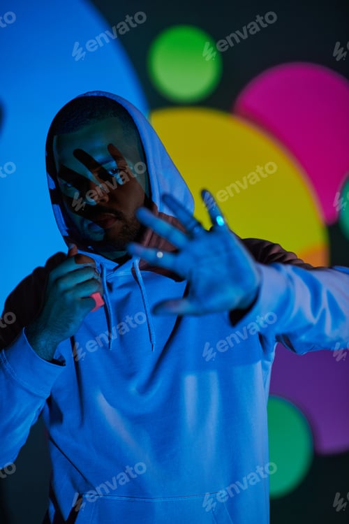 Preview: good looking man with hood on putting hand in front of camera in projector lights, fashion concept