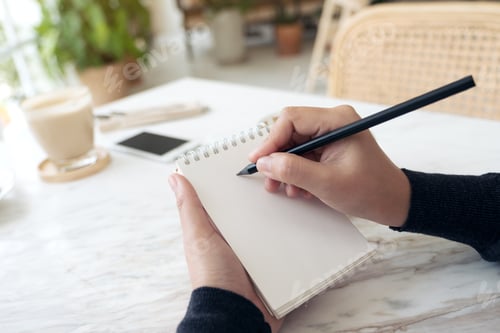 Preview: a hand writing down on a white blank notebook with coffee cup and dessert on table in cafe