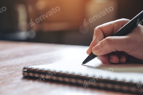 Preview: Closeup image of a hand writing down on a white blank notebook on wooden table