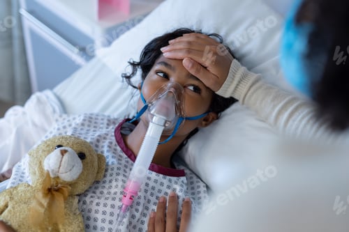 Preview: Mixed race mother in face mask touching sick daughter's forehead on oxygen ventilator in hospital