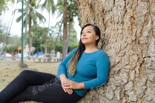 Preview: Woman seated leaning against a tree with pensive attitude.