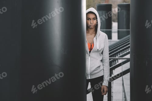 Preview: Attractive young sportswoman in hood posing on stadium and looking at camera