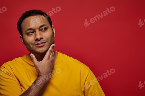Preview: dreamy indian man in yellow t-shirt touching chin and looking away on red background, thoughtful