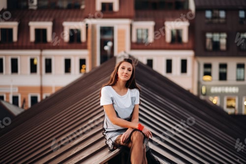 Preview: A girl sits on the roof of a house in the city in the evening. Portrait of a model in a dress and