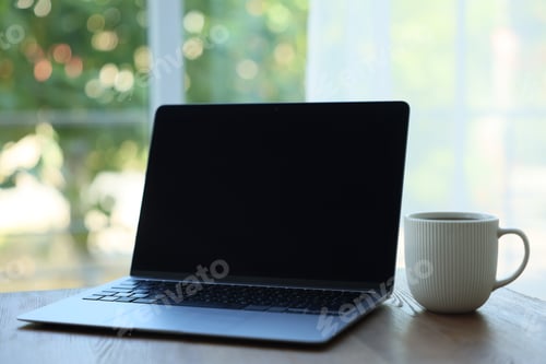 Preview: Open laptop and cup on wooden table against blurred green background, closeup. Mockup for design