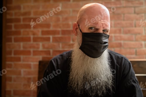 Preview: Serious man with long gray beard wearing face mask to protect from corona virus Covid 19