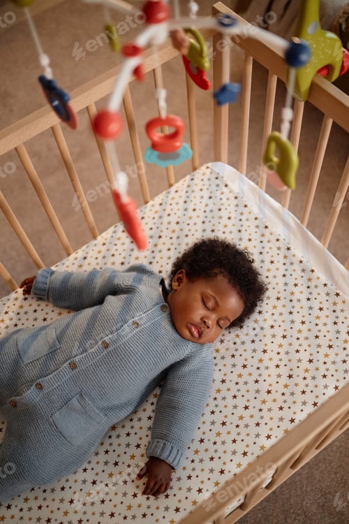 Preview: Sleeping Black Girl Lying in Baby Crib with Mobiles Hanging above Child Indoors
