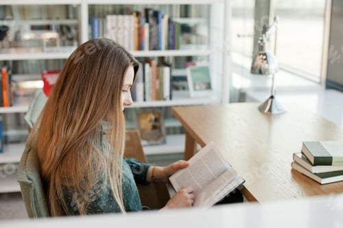 Preview: Student studying at a public library: reading a book at a desk with lots of books on bookshelves
