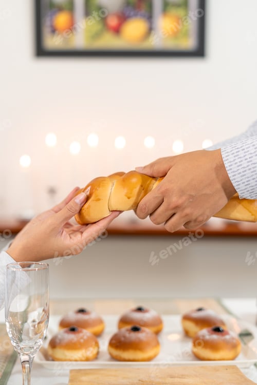 Preview: Vertical photo of two hands pass challah bread during a Hanukkah meal.