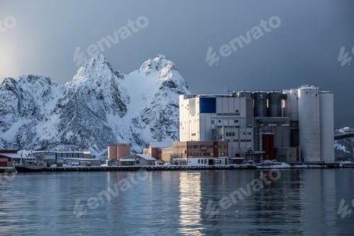 Preview: Industrial buildings against a backdrop of snowy mountains reflected on calm water at dawn