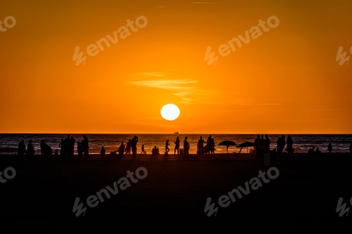 Preview: Silhouette of a beach full of people under the cloudless red sunset sky reflected on the sea