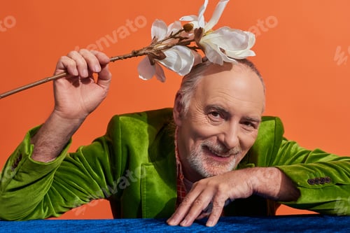 Preview: happy aging concept, delighted and bearded senior man in green velvet blazer sitting at table