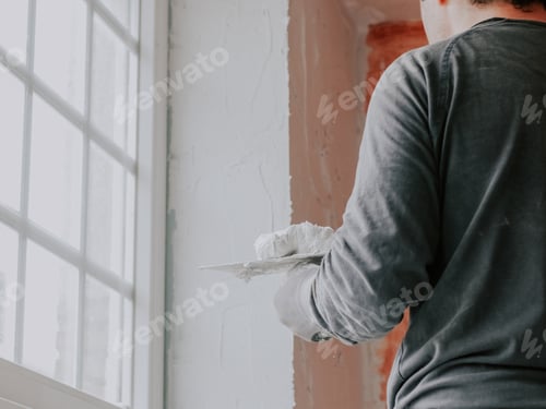 Preview: A young caucasian guy applies fresh putty with a small spatula to a window opening near the ceiling,