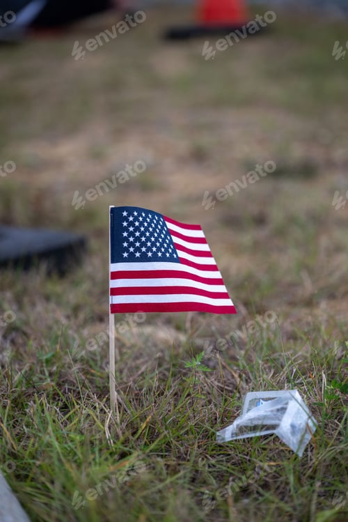 Preview: Vertical closeup of a small United States flag on the grass with a piece of garbage on the side