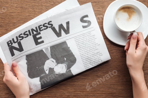 Preview: cropped image of journalist holding business newspaper and cup of coffee at wooden table