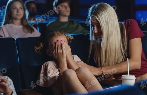 Preview: Worried small girl covering eyes in the cinema while watching film, mother comforting her