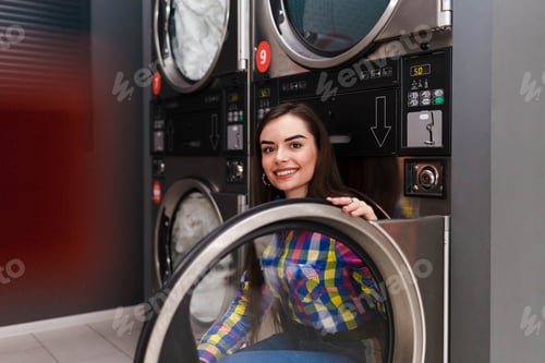 Preview: Positive young woman sitting on the dryer machine in the self-service laundromat