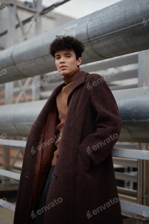 Preview: Fashionable young man in brown coat and sweater leaning against metal railing with large pipe in
