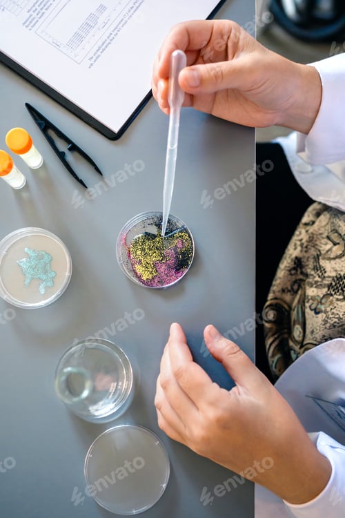 Preview: Female chemist adding drop of liquid from from pipette over petri dish with mix of glitters in lab