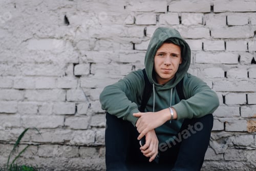 Vista previa: Joven en sudadera con capucha sentado junto a la pared de ladrillo