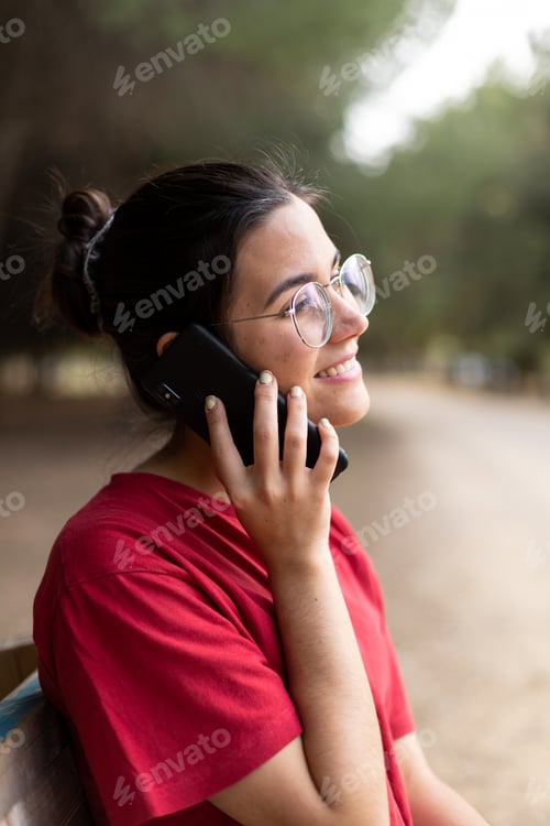 Preview: Young attractive woman talking through the phone and smiling in a park, sit down in a bench
