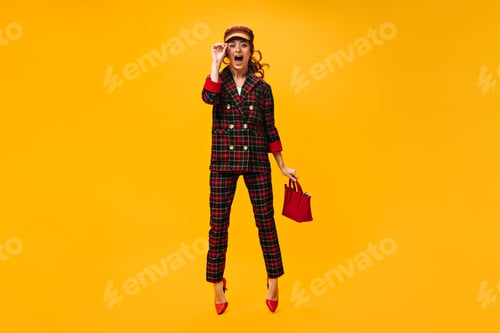 Preview: Full length woman in plaid outfit jumps on orange background and holds handbag. Charming girl in ey