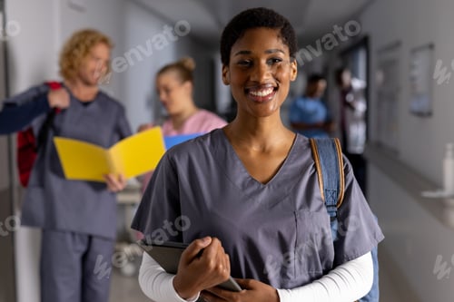 Preview: Portrait of happy african american female doctor wearing scrubs with tablet in corridor at hospital