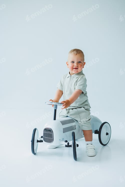 Preview: Stylish one-year-old boy in a suit with shorts and a T-shirt rides a car. Toddler boy on a white