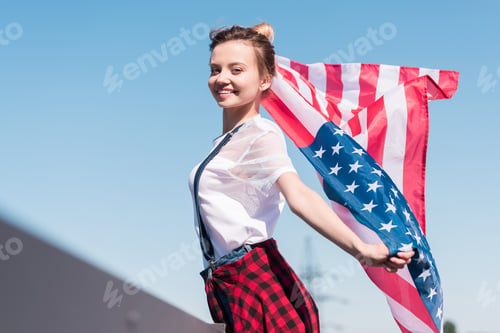 Preview: smiling young woman holding american flag against blue sky, independence day concept