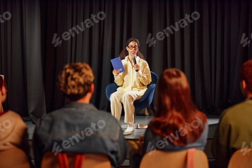 Preview: Young Adult Woman Hosting Public Reading Event on Stage Facing Audience