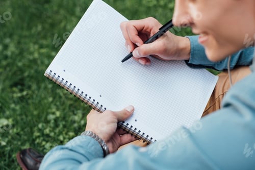 Preview: cropped shot of smiling young man writing in blank notebook while sitting on grass