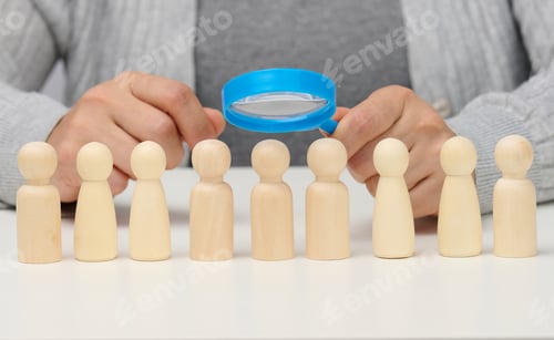 Preview: Figurines of men on a white table, a female hand holds a magnifying glass over one.