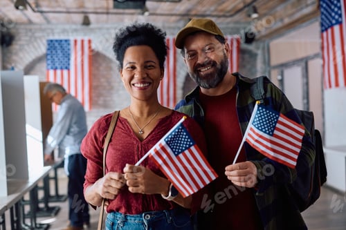 Preview: Happy multiracial couple with US national flags at polling place looking at camera.