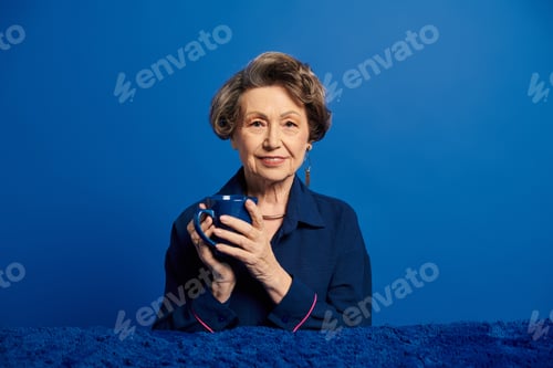 Preview: A beautiful woman in her 60s showcases her style while holding a cup against a blue backdrop.